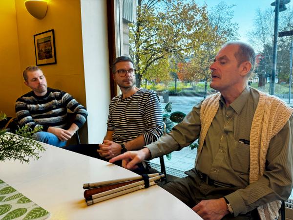 Mats Nilsson sitter vid ett bord. Framför honom ligger den vita käppen. Mats har kort mörkt hår och bär en skjorta i olivgrönt med smala ränder. Ovan på har han en stickad väst i offwhite.Brevid honom sitter två politiker.