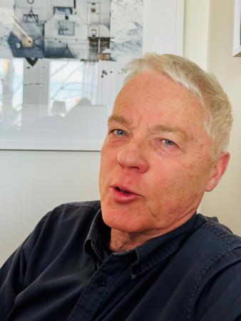 Mats har kort, grått hår och ser engagerad ut. Han har en mörkblå tröja på sig och sitter framför en tavla med ett kollage av vita detaljer från olika hus.