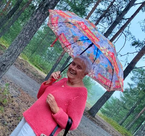 Inger står ute bland tallar hon har en rosa, stickad tröja och håller i ett paraply. Hon har kort grått hår och ser glad ut.