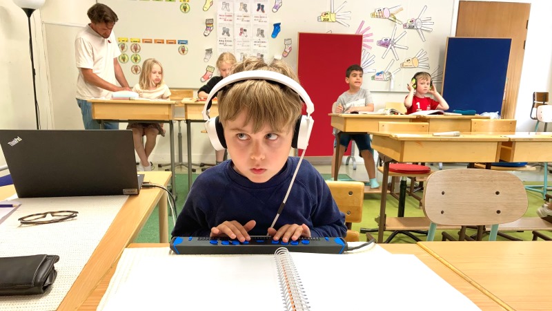 Walter sitter i klassrummet med hörlurar på. Han skriver på sin punktdisplay och ser koncentrerad ut. Han har mellanblont hår och bär en blårandig tröja.