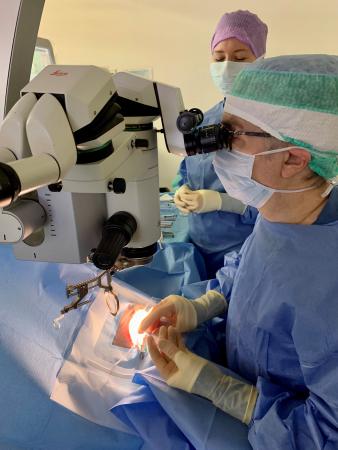 Daniel tittar in i ett mikroskop och ger en patient en bedövningsspruta i ögat. Han har munskydd, handskar, hårskydd och blå kläder. Bakom honom står en operationssköterska.
