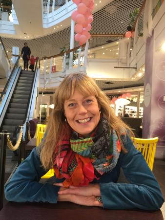 Ann-Sofie Sköld Åberg bär en petrolgröntröja och en mångfärgad sjal i röda nyanser. Hon har långt, ljust hår och skrattar på bilden. Hon svilar armarna på ett bord i ett köpcentrum och man ser en rulltrappa bakom henne till vänster, ovanför henne hänger rosa ballonger och man skymtar knallgula stolar i bakgrunden.