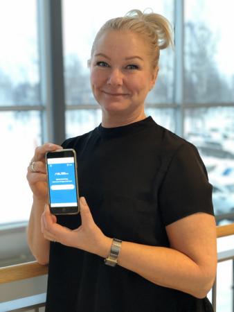 Lisette Sällström visar upp en telefon med inloggningsappen till Region Norrbotten. Lisette har långt, ljust hår uppsatt i en svans. Hon har en svart, kortärmad tröja på sig.