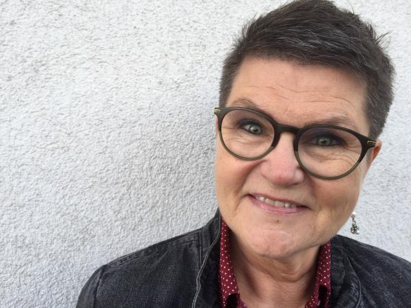 Agneta titta rakt in i kameran och ler. Hon har kortklippt mörkt hår, glasögon med mörka bågar  och pärlörhängen. Hon har en röd skjorta med vita prickar och en mörk jeansjacka.