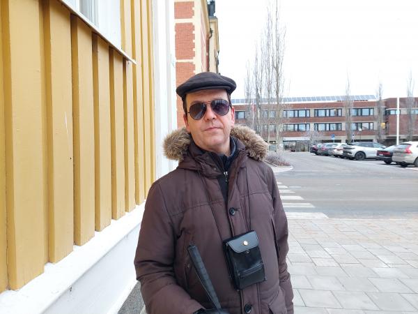 Matti Jatkola är lärare på Sverigefinska folkhögskolan i Haparanda. På bilden står han utanför ett gult hus i centrala Haparanda. Han har en basker på huvudet och solglasögon på sig. Runt halsen hänger en väska med hans mobiltelefon. Han har vinterjacka.