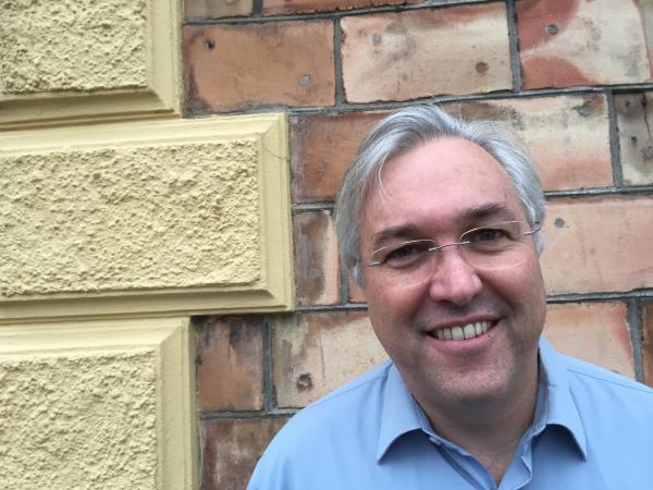 Greg Olsson Lalore står mot en gul/rostfärgad tegelvägg. Han är klädd i en ljusblå skjorta och har grått, kort hår. Han har glasögon och skrattar på bilden.