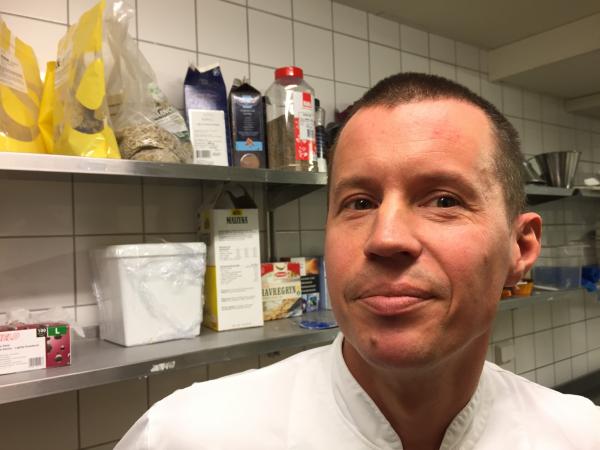 Benny står i sitt bageri iförd en vit rock. Han har kort mörkt hår. Bakom honom skymtar olika ingredienser på hyllor.