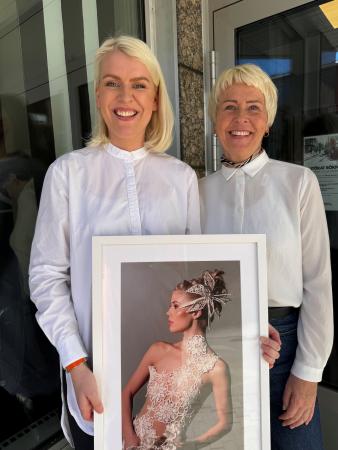 Olivia är längre än sin mamma Anneli. Båda bär vita skjortor och är blonda. Oliva med en axellång page och Anneli har en kor uppklippt frisyr med kortare nacke och flikar fram vid öronen. De ser glada ut och Olivia håller en bild på årets brud som hon vunnit pris med.