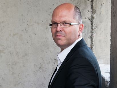 Daniel är snaggad och bär glasögon. Han är klädd i kostym och vit skjorta. 