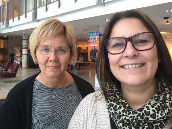 Åsa har blont kort hår och glasögon. Hon bär en grå tröja och svart kofta. Pia har axellångt mörkt hår och glasögon med mörk båge. Hon har en vit tröja och en leopardfärgad sjal.
