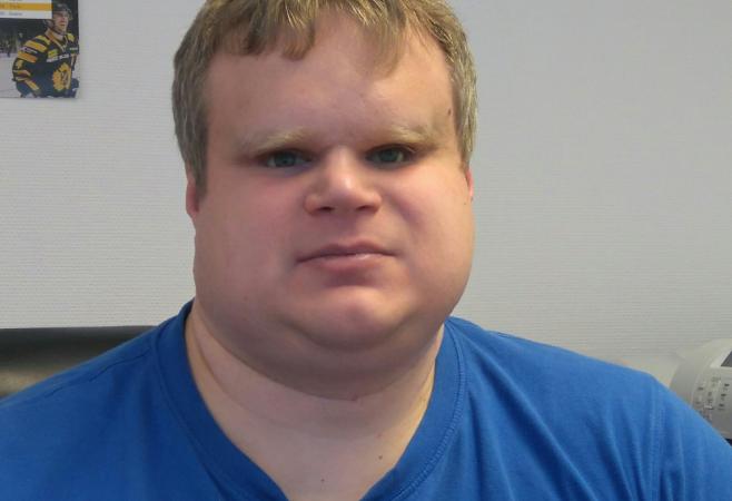 David sitter i en stol vid sitt skrivbord. Han har kort, ljust hår och bär en klarblå t-shirt