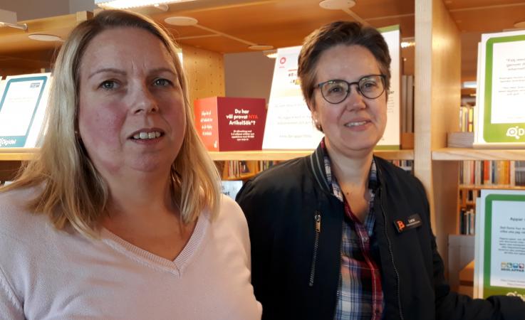 Ingela Karlsson Broström och Lena Vesterlund står i stadsbibliotekets digidelcenter. Ingela har långt ljust hår och är klädd i en ljusrosa tröja. Lena har kortklippt mörkt hår och glasögon. Hon har en mörk jacka utanpå en rutig skjorta.
