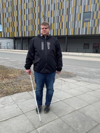 David står framför ishockeyhallen i Skellefteå. Inte oväntat är den dekorerad i svart ocg gult. David har blå jeans och jacka. Han håller i en vit käpp och är blond med lockigt hår.