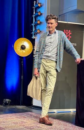 Gustav är lång och smal. Han har mörkt, mycket lockigt hår och bär en kofta över skjorta.