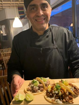 Kocken Homero håller fram en träbricka med tacos. Han har en svart kockjacka och mössa. Han ser glad ut.