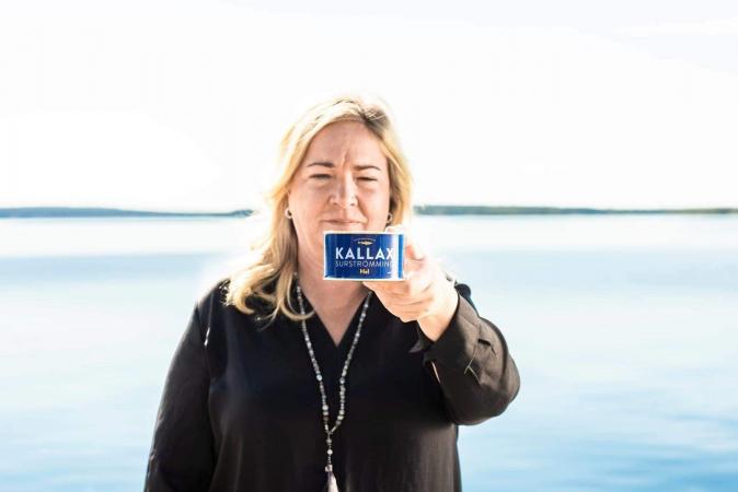 Veronica står framför vatten och håller i en surströmmingsburk. Hon har långt ljust hår och bär en svart jacka och ett långt halsband.