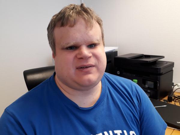 David sitter i en stol vid sitt skrivbord. Han har kort, ljust hår och bär en klarblå t-shirt