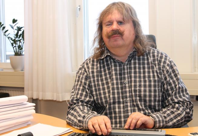 Håkan Thomsson sitter vid ett skrivbord. Han har långt brunt hår och stor mustasch. Han är klädd i en brun och vitrutig skjorta. 