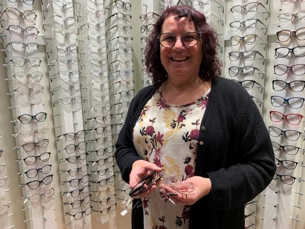 Karin har mörkt, lockigt hår och bär mönstrade kattögonformade glasögon. Hon står framför en vägg av bågar och håller ett par i händerna.