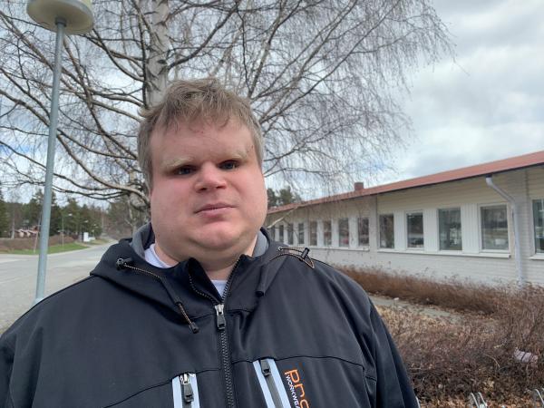 David har kort ljust hår. Han har en blå jacka och står framför ett träd.