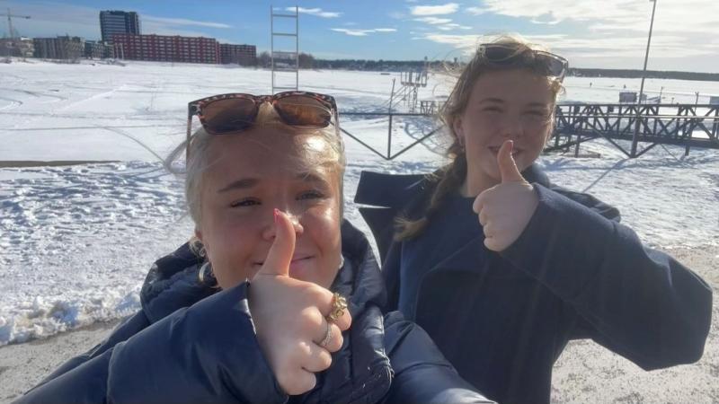Tuva-Li är blond och har ett par solglasögon på huvudet. Thelma har mörkt, långt hår. De står ute på isvägen framför Luleå-skylten och gör tummen upp. Kanske till sin praktik på taltidningen i veckan?