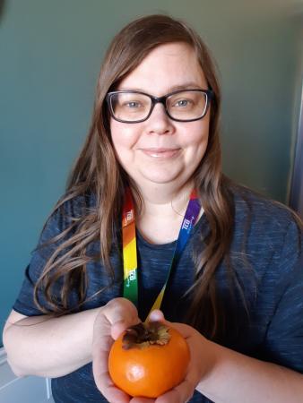 Emma som håller i en orange sharonfrukt har långt mörkt hår och glasögon. Hon ser glad ut på bilden.