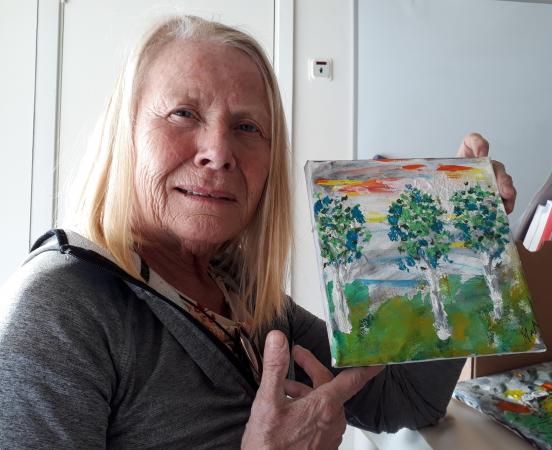 Marianne Nordqvist håller upp en tavla. Hon har ljust halvlångt hår och har på sig en grå tröja.