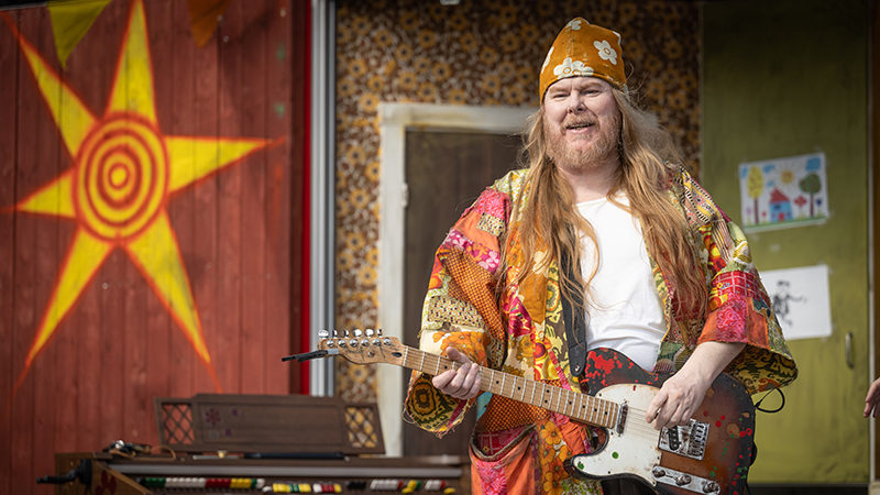 Mattias har långt, rött hår. Han har en färggrann tehuva på huvudet och en orange morgonrock. På bilden sjunger han och spelar gitarr i huvudrollen som Loranga.