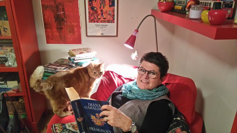 Agneta sitter i en röd fåtölj i ett rum inrett i rött. Hon håller i en bok och ser glad ut.