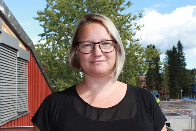 Mascha Paulsen står ute och man ser en röd byggnad på vänster sida.Bakom henne en grönskande björk. Mascha har en svart t-shirt och bär glasögon. Hon har en blond page som är lite längre på ena sidan. Hon ser glad ut.