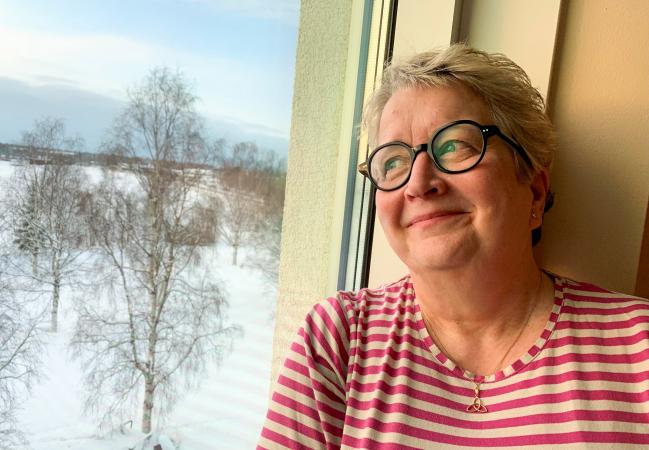 Gunilla har kort hår och glasögon med mörka bågar. Hon bär en röd- och vitrandig tröja och tittar drömskt ut genom fönstret.
