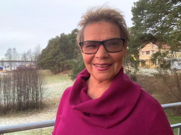 Ulla har kort melanblont hår och glasögon med mörk båge. Hon har en lila stickad tröja med krage. Hon ser glad ut.