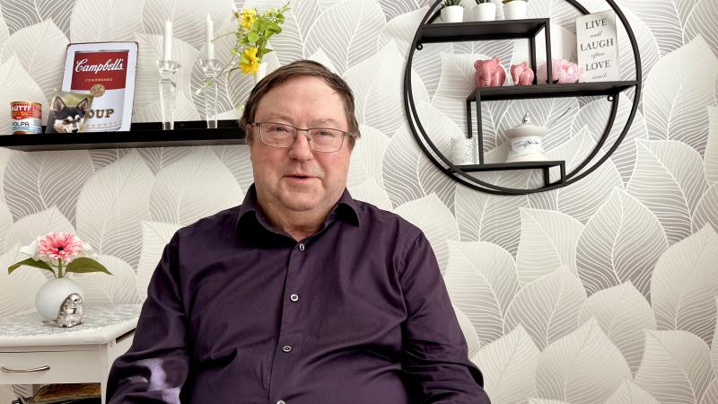 Roland har vintröd tröja och står framför en vägg med tapet som har stora vita blomblad. Roland har mörkt kört hår och bär glasögon. Han ser glad ut!