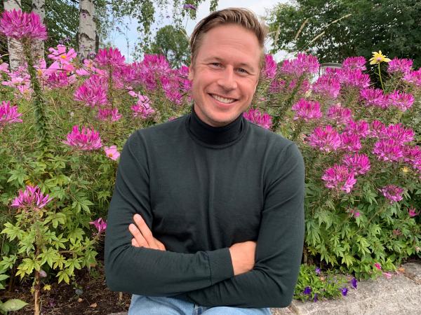 Viktor har en grå tröja och sitter framför ett hav med höga rosa blommor. Han har armarna i kors och hans mellanblonda hår är kammat bakår. Han skrattar.