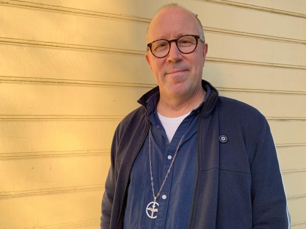 Anders har kort hår och bär glasögon med mörka bågar. Han är klädd i mörkblått och har symbolen för diakoni i en lång kedja runt halsen: en rund cirkel med ett kors där en duva med myrtelkvist i näbben landat.