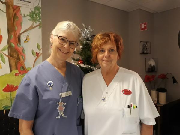 Christina har en blå skjorta och vitt hår. Hon bär glasögon. Anette har en vit sjuksköterskeskjorta och hennes hår är rött i en kort frisyr.