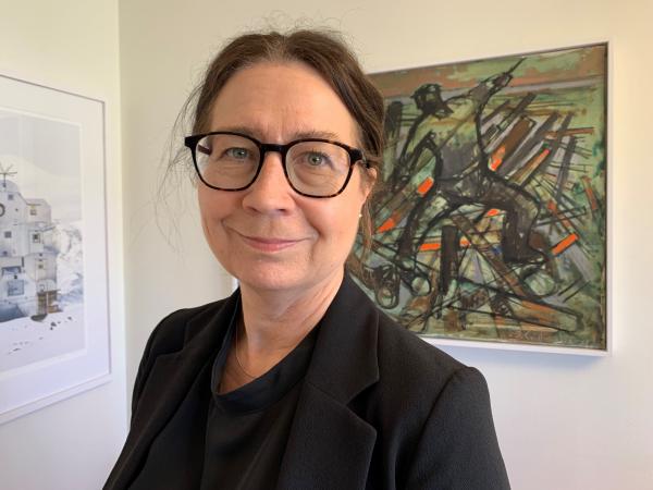 Liselott har mörkt uppsatt hår och glasögon med mörka bågar. Hon har en svart skjorta och ser glad ut. Hon står framför en tavla målad av Ingemar Callenberg.