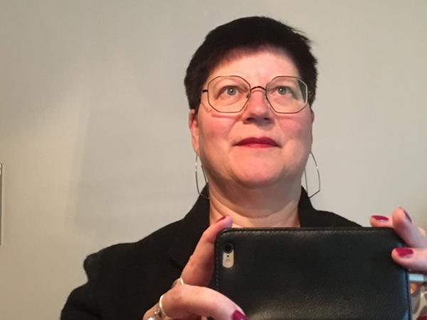 Susanne Jonsson tar en klassisk selfie med mobilen. Dvs hon fotar sig själv i en spegel. Något som många gör 2018. Susann har mörkt kortklippt hår. Hon bär glasögon och har rött läppstift. Fingrarna som håller i mobilen är knallröda. Hon har en svart jacka. På bilden ser hon finurlig ut och blickar upp mot högra hörnet.