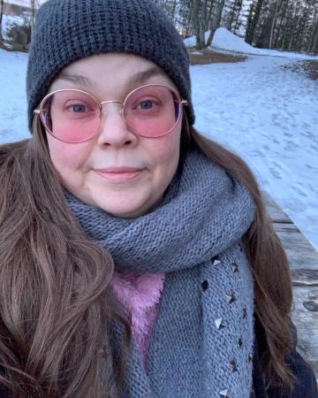 Emma har långt mörkt hår under en grå toppluva. Hon har runda glasögon med rosa toning.