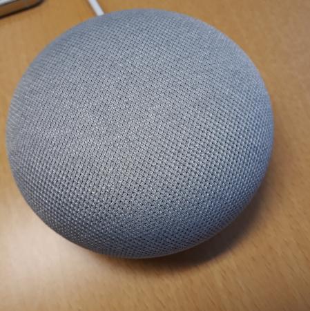 Bilden visar en Google assistent - en rund, grå högtalare som är cirka tio centimeter i diameter. Den ligger på ett träfärgat bord.