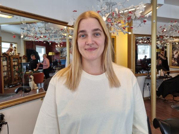 Erica Lindkvist står inne på frisörsalongen. Hon har en vit tröja. Långt, blont och rakt hår. Markerade ögonbryn och en ring i näsan. I bakgrunden skymtar speglar och andra frisörer som jobbar.