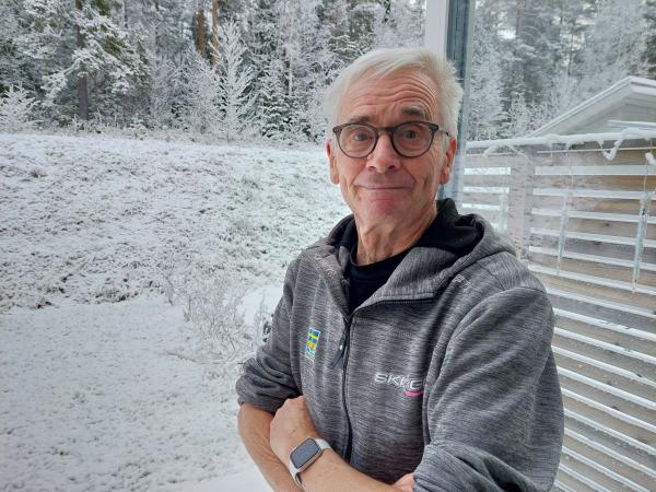 Sven-Åke Lundbäck står ute på sin altan. Det är snö i bakgrunden. Han har en grå tröja och armarna i kors. Han har glasögon och ett litet leende.