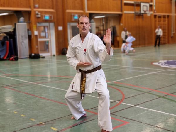 Mats är klädd i vit karatedräkt med ett brunt bälte. Han står redo att fightas. Mats har väldigt lite, grått hår och ser bestämd ut.