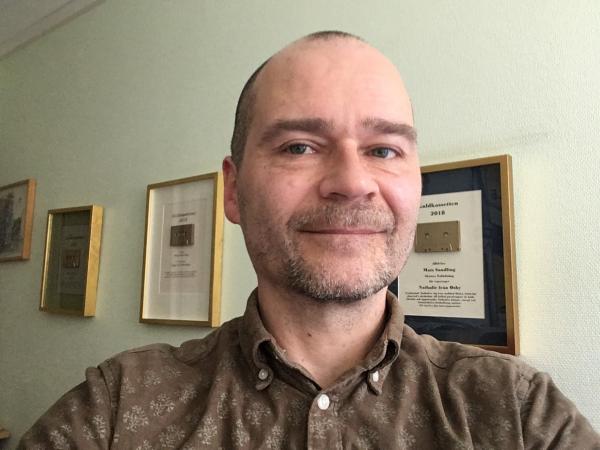 Mats har kort brunt hår och mustasch skägg. Han ser glad ut och står framför guldkassettvinnartavlor