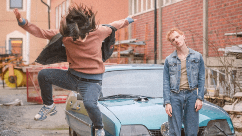En blond kille i blå jeansjacka och jeans står framför en ljusblå bil. En annan person gör ett hopp med armarna utslagna.