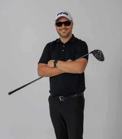 Torbjörn har coola svarta solglasögon och bär en vit keps. Han har skäggstubb och ser glad ut. Han har en svart pikétröja och svarta byxor. Han håller i en golfklubba