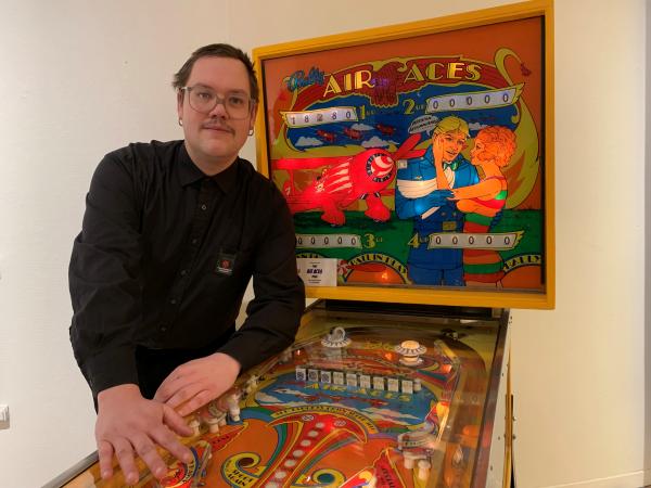 Andreas har kort hår och glasögon. Han står lutad på ett flipperspel i orange och gult.