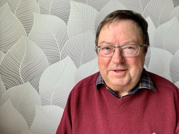 Roland har vintröd tröja och står framför en vägg med tapet som har stora vita blomblad. Roland har mörkt kört hår och bär glasögon. Han ser glad ut!