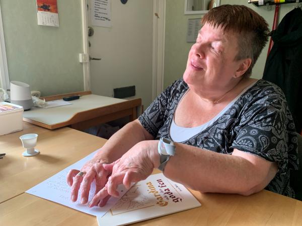 Anita har kort grått hår och på bilden läser hon sin barnbok Gardina och en goda fen i punktskrift. Ett exemplar av boken syns på bilden.