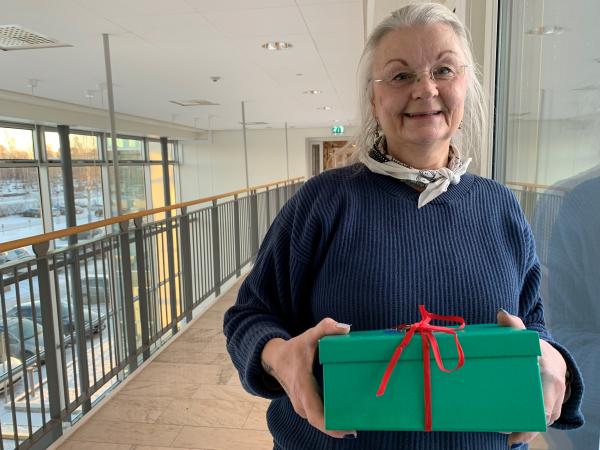 Agneta har vitt hår och glasögon med tunnbåge.Hon ser glad ut och håller i en grön låda med ett rött band.